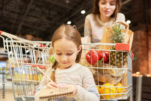 Warm-toned portrait of family doing grocery shopping in supermarket, focus on little girl holding shopping list standing by cart photo