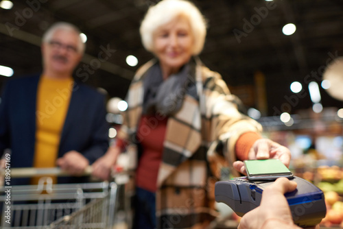 Portrait of modern senior couple buying groceries in supermarket paying with NFC payment via smartphone, focus on foreground, copy space