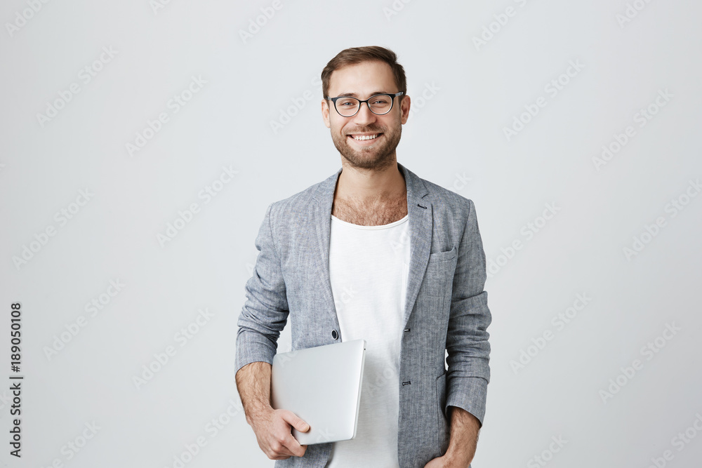 Horizontal portrait of glad male office worker with thick beard, wearing spectacles, holding laptop in his hands, smiles with perfect teeth at camera. Occupation and modern technologies concept Horizontal portrait of glad male office worker with thick beard, wearing spectacles, holding laptop in his hands, smiles with perfect teeth at camera. Occupation and modern technologies concept