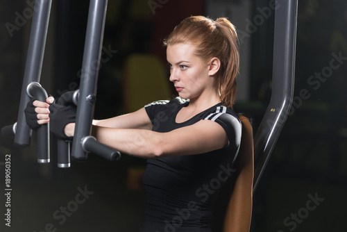 Woman at the sport gym doing arms exercises on a machine. Dark sport club.