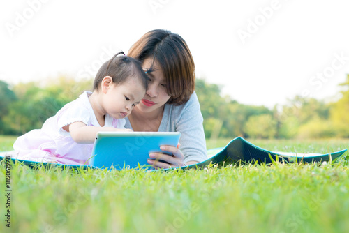 Mother and daughter outdoors in a meadow playing tablet. Mother with her daughter lying on grass and using digital tablet together. Mother and daughter education concept.