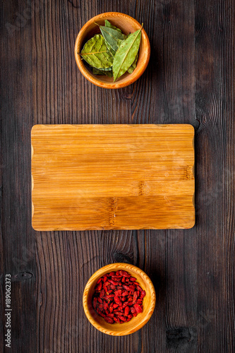 Make menu or write recipe. Mock up for menu or recipe. Wooden cutting board near ingredients in bowls bay leaf and godji on dark wooden background top view