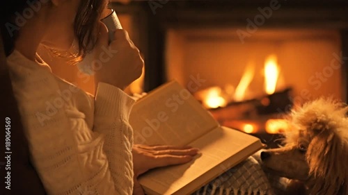 Young woman with her dog sitting at home by the fireplace and reading a book.