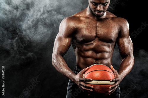 Portrait of afro-american sportsman, basketball player with a ball over black background. Fit young man in sportswear holding ball. © Mike Orlov