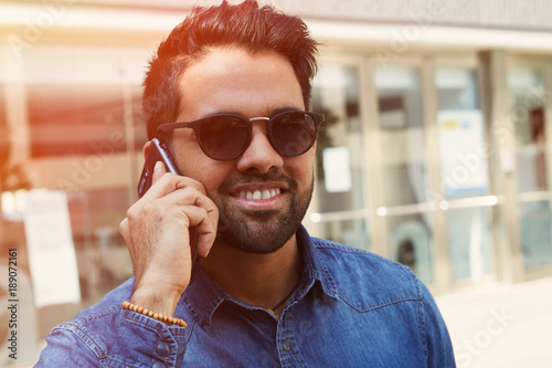 Attractive young man with cell smiling guy relaxing outdoors making mobile call, successful confident hipster having pleasant phone conversation. photo