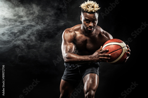 Portrait of afro-american sportsman, basketball player with a ball over black background. Fit young man in sportswear holding ball. © Mike Orlov