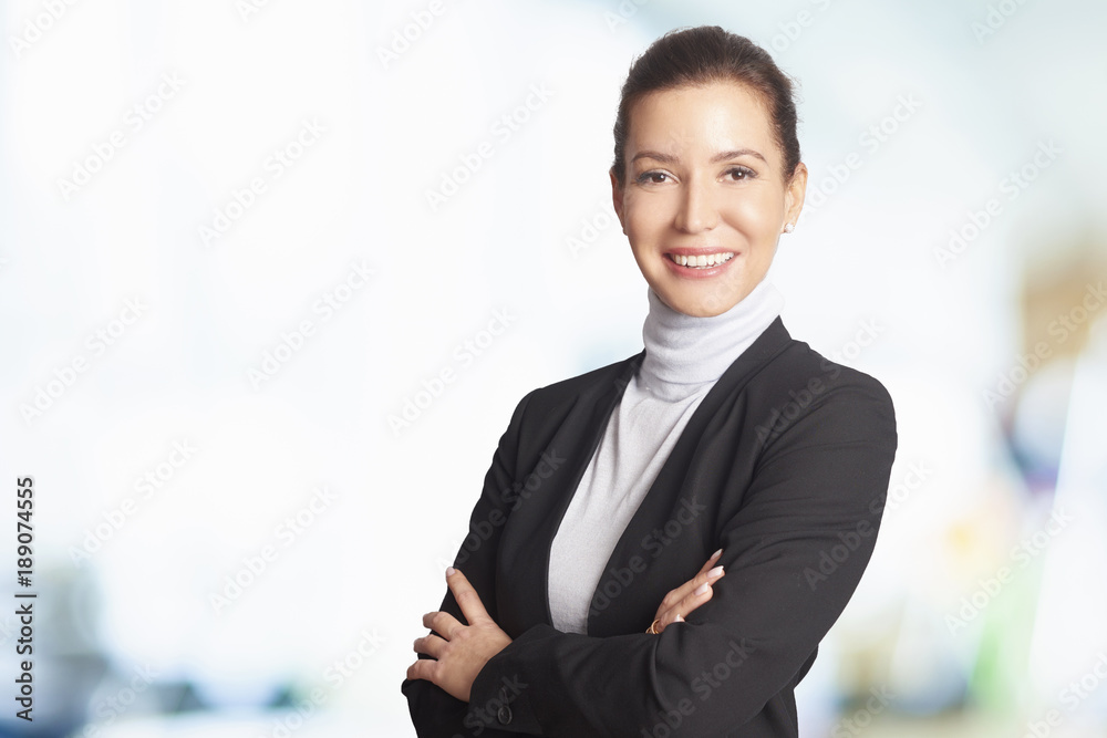 Businesswoman at the office. Smiling middle aged woman standing at the office and looking at camera while her female colleague standing in the background. Businesswoman at the office. Smiling middle aged woman standing at the office and looking at camera while her female colleague standing in the background.