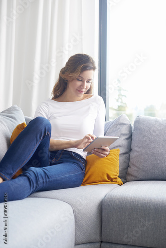 Woman browsing on the internet. Smiling middle aged woman using digital tablet while relaxing on sofa at home.