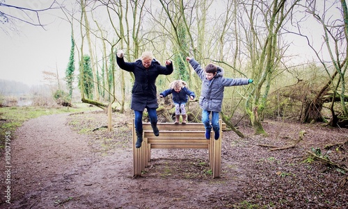 Mother and two children jumping off a wooden steps in forest, Kingsbury, Warwickshire, England, UK photo