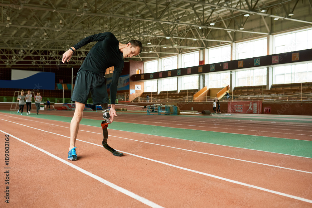 Full length portrait of young handicapped athlete with artificial foot warming up before running practice in modern gym stretching to sides, copy space
