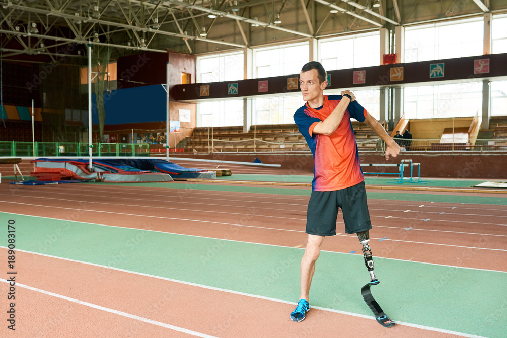 Fototapeta premium Full length portrait of young amputee sportsman warming up before running practice in modern indoor stadium , copy space