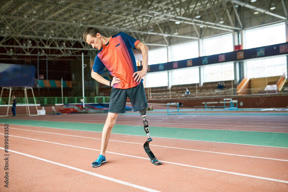 Full length portrait of young handicapped sportsman with artificial foot warming up before running practice in modern gym stretching to sides, copy space