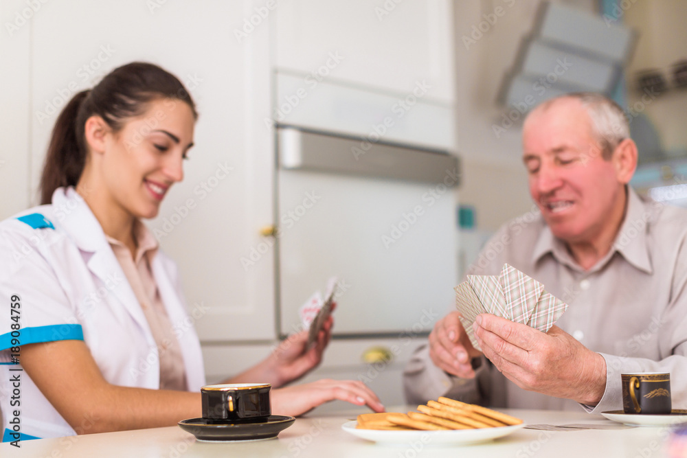 Obraz premium Portrait of the old joyful man and his young cheerful doctor who sitting at the table in the kitchen and they playing cards