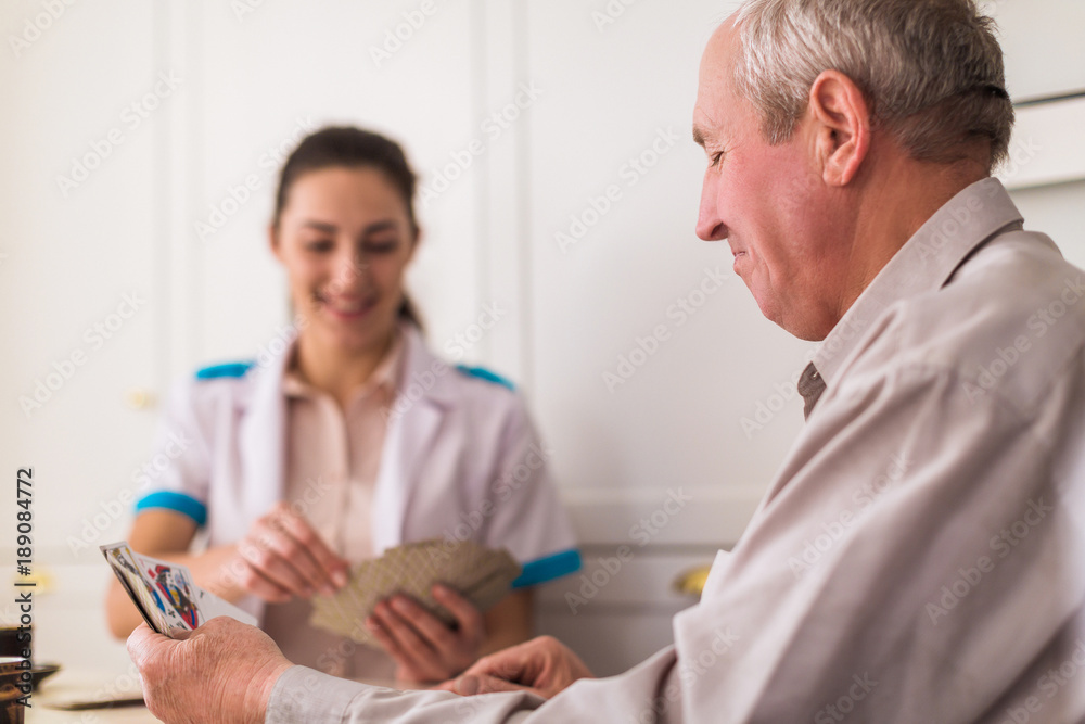 Fototapeta premium Side view of the old joyful man who sitting at the table with his young cheerful doctor in the kitchen and they playing cards