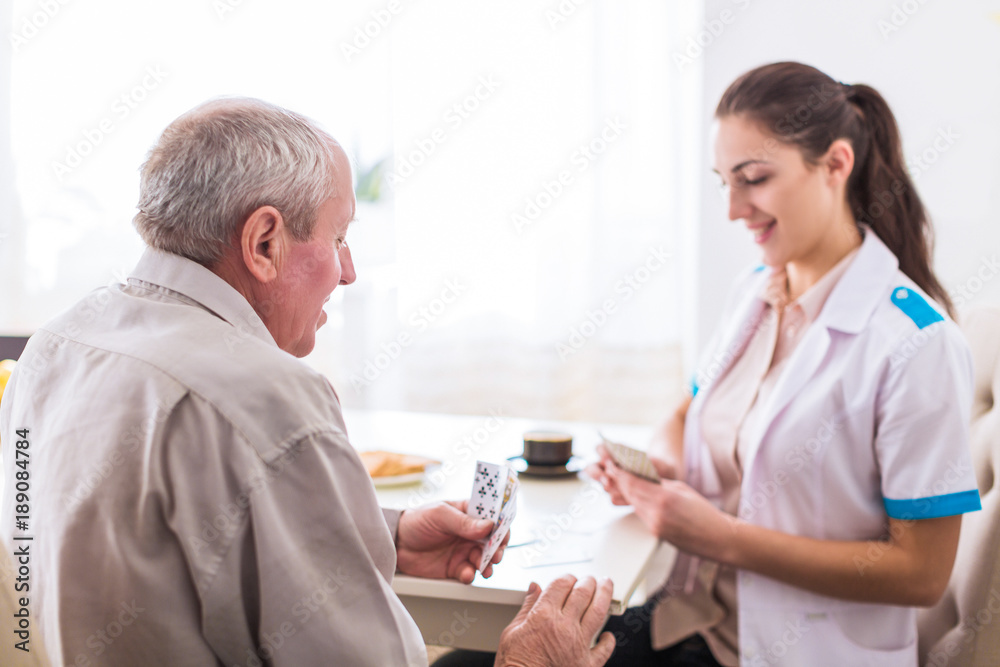 Obraz premium The young happy nurse sitting at the table with the old joyful man in the kitchen and they playing cards