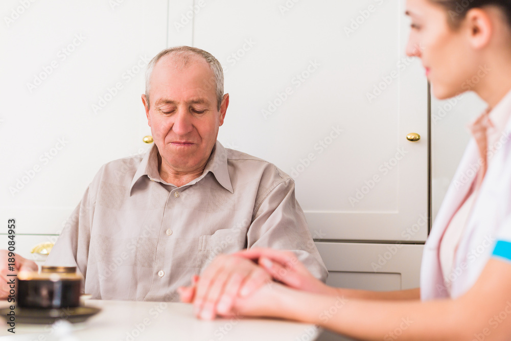 The old ill man sitting at the table in the kitchen and the young nurse sitting near him and holding his hand The old ill man sitting at the table in the kitchen and the young nurse sitting near him and holding his hand
