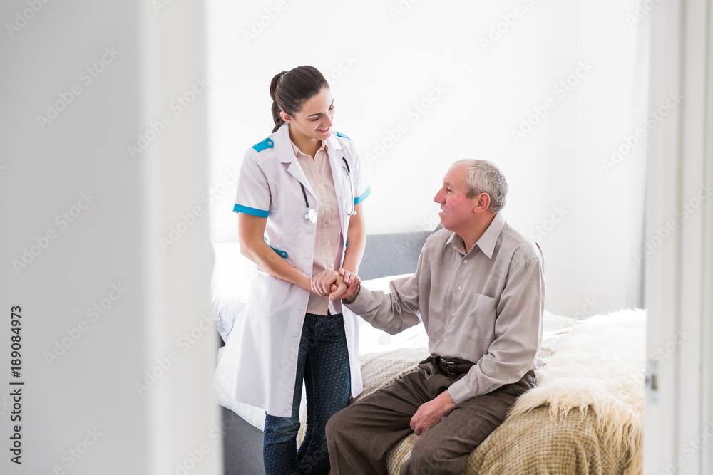 Obraz premium The old ill man sitting on a bed in the room at home and the young smiling nurse standing near him and talking with him