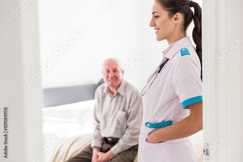 The old ill man sitting on a bed in the room at home and the young smiling nurse standing in the room near the door