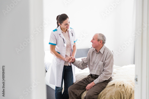 The old ill man sitting on a bed in the room at home and the young smiling nurse standing near him and talking with him