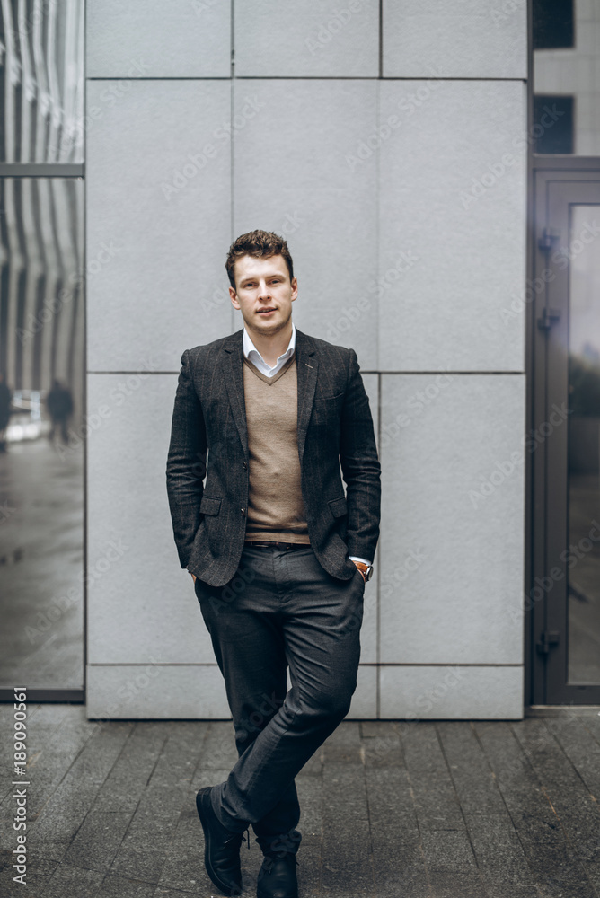 Photo of a stylish young man who stands outdoor against the background of a gray building and smiling Photo of a stylish young man who stands outdoor against the background of a gray building and smiling