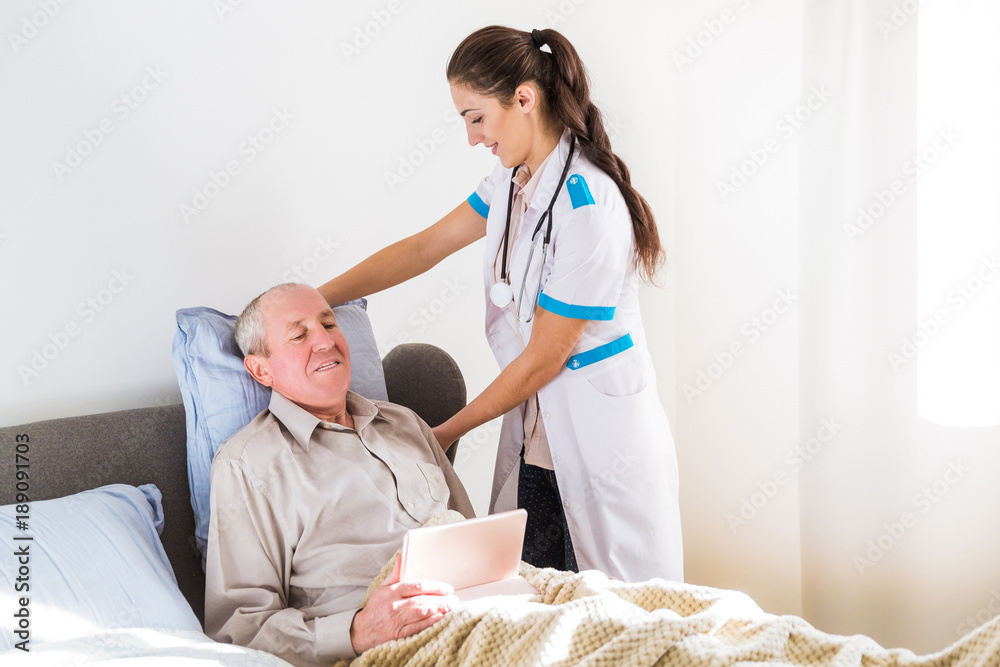 Obraz premium The old, ill, man lying on a bed and looking at the tablet and the young doctor standing near him and fixing a pillow to him