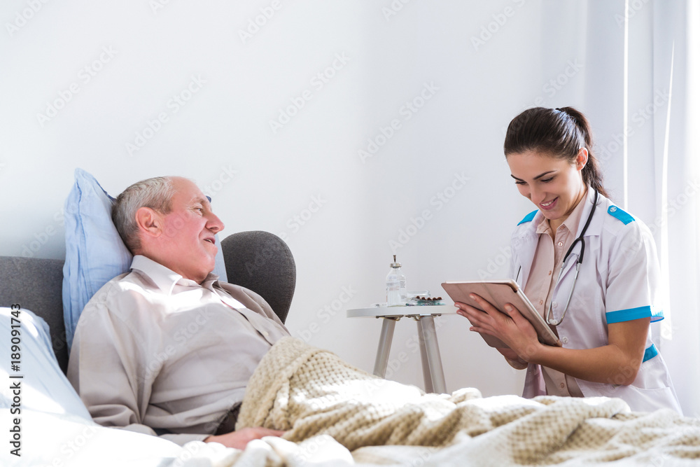 The old, sick, man lying on a bed in the room looking at the young doctor who is sitting near him and looking at the tablet The old, sick, man lying on a bed in the room looking at the young doctor who is sitting near him and looking at the tablet