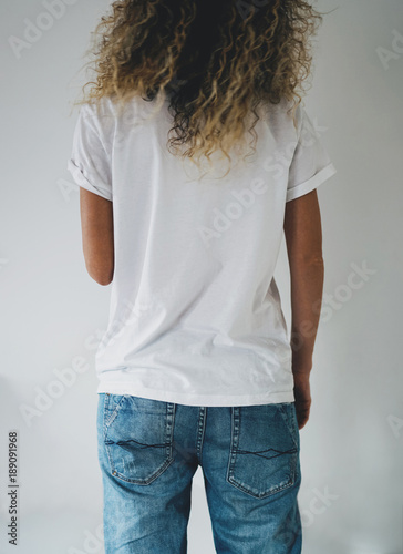 An attractive young woman dressed in a white blank t-shirt and blue jeans posing on a background of a white wall. Vertical mock up. empty space for you logo or design. Back view.