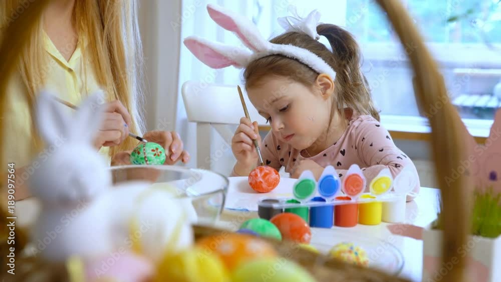 Happy Easter! Young mother and her cute little daughter wearing funny rabbit ears are coloring easter eggs sitting at a festive table with basket and Bunny Happy Easter! Young mother and her cute little daughter wearing funny rabbit ears are coloring easter eggs sitting at a festive table with basket and Bunny
