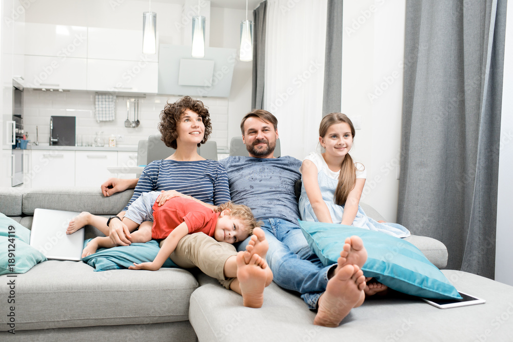 Happy family of four sitting on cozy sofa and watching their favorite TV program, interior of modern studio apartment on background