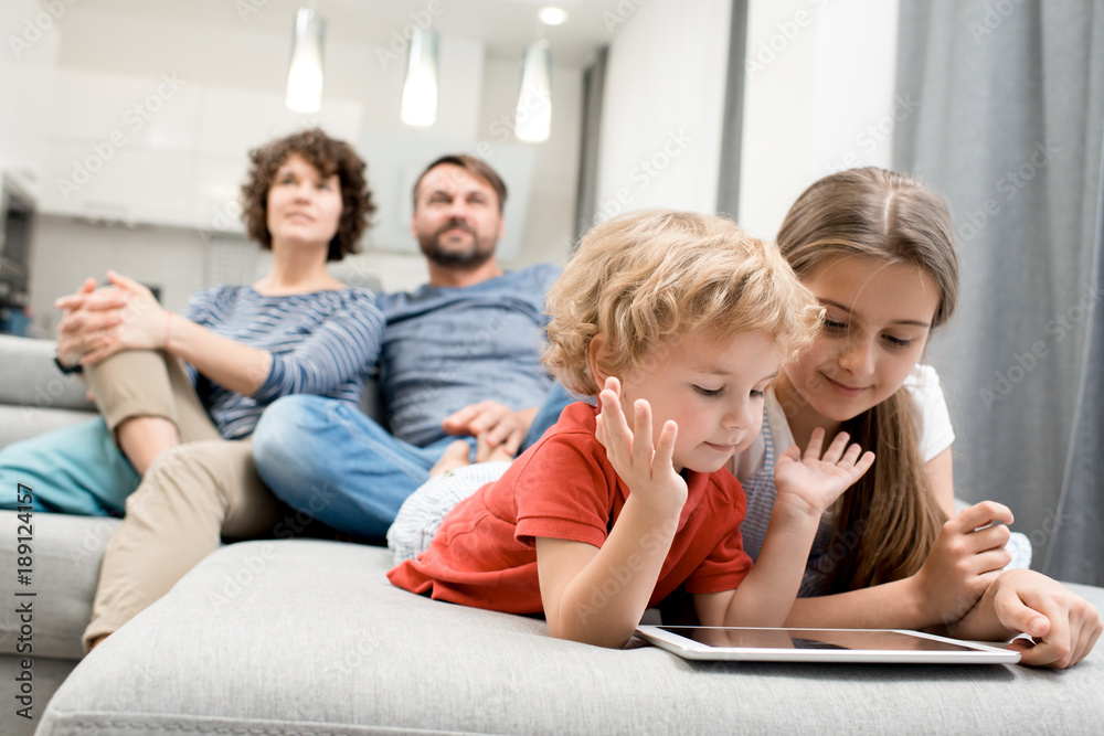Pretty little girl and her curly brother lying on cozy couch and playing game on digital tablet while their parents wrapped up in watching TV, interior of studio apartment on background Pretty little girl and her curly brother lying on cozy couch and playing game on digital tablet while their parents wrapped up in watching TV, interior of studio apartment on background