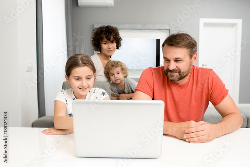 Smiling family of four gathered together in studio apartment and watching their favorite cartoons on laptop