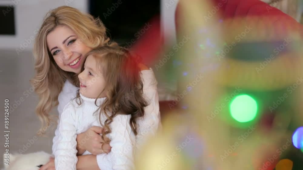 Happy mother and little daughter sit in an embrace near the Christmas tree, next to them goes a large fluffy white dog. Happy mother and little daughter sit in an embrace near the Christmas tree, next to them goes a large fluffy white dog.