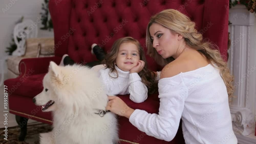 Young mother and little daughter playing with a white husky Samoyed sitting on the beautiful red couch. Young mother and little daughter playing with a white husky Samoyed sitting on the beautiful red couch.