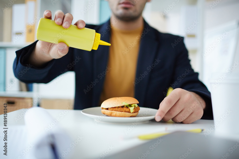Office manager adding mustard from plastic bottle to his hamburger before eating it Office manager adding mustard from plastic bottle to his hamburger before eating it