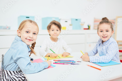 Happy little girls and boy looking at camera at drawing lesson in kindergarten