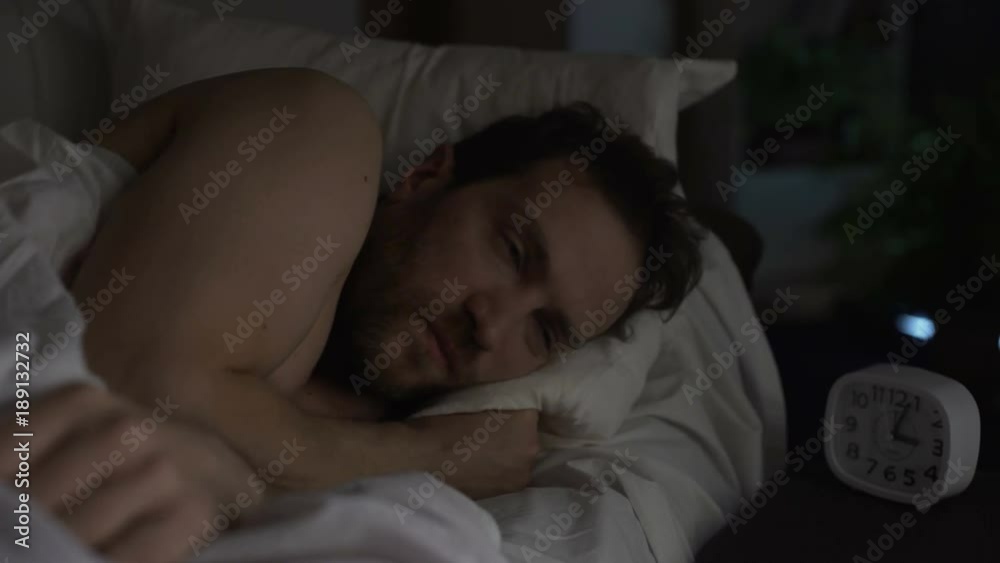 Guy turning in bed unable to fall asleep, looking angrily at clock, insomnia