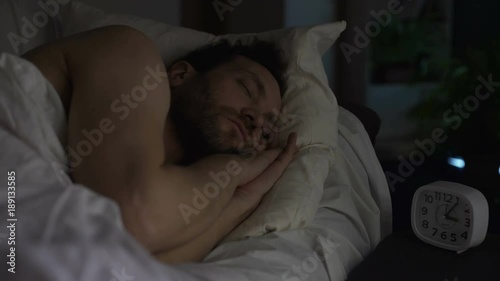 Bearded man sleeping on sofa bed, clock standing on night table, late night rest photo