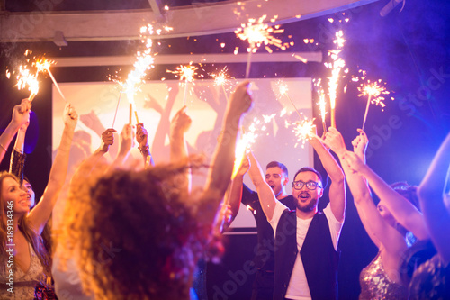 Crowd of trendy young people holding glittering sparklers dancing in night club celebrating holiday
