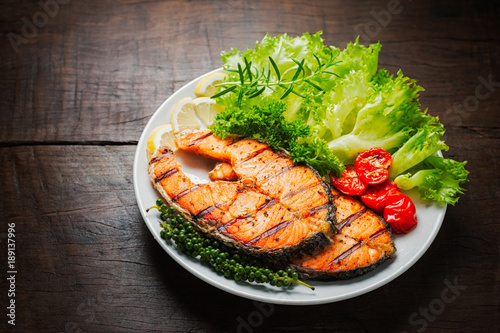Grilled salmon steak served with grilled tomatoes, salad, lemon sliced, pepper seeds, rosemary, parsley on white plate on wooden table.