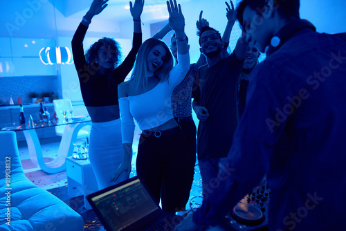Group of modern young people dancing listening to DJ playing music at private house party, lit by blue light