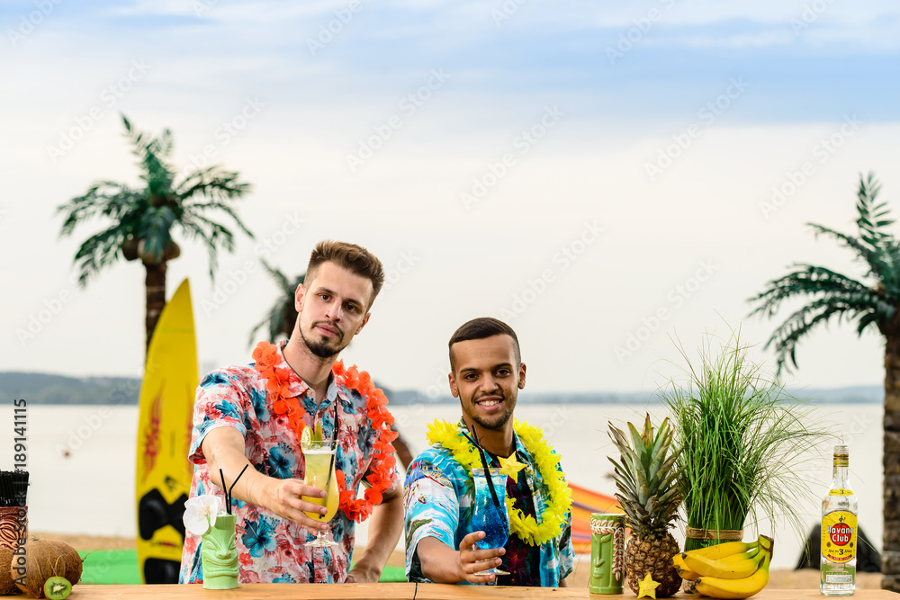 Handsome caucasian bartender and Latin American barman standing near the bar counter and holding cocktails on the ocean. Concept of preparation, relaxing and fun