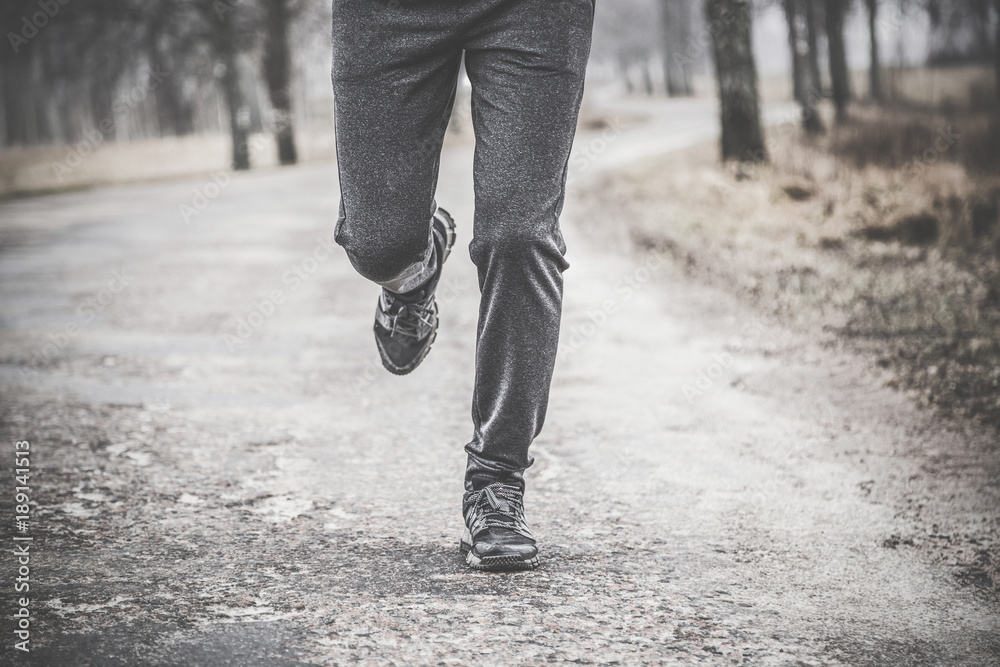 Young man running away on road outside of city in the dark cloudy afternoon. Daily outdoor active lifestyle. Enjoying sport concept. Runner's legs with sneakers. Young man running away on road outside of city in the dark cloudy afternoon. Daily outdoor active lifestyle. Enjoying sport concept. Runner's legs with sneakers.