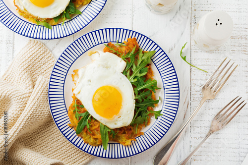 Fried egg with potato pancake, arugula and avocado on ceramic plate for breakfast on white wooden table background. Selective focus. Top view. Copy space.