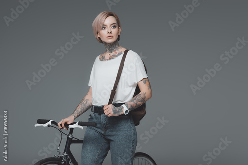 beautiful tattooed girl with backpack standing with bicycle and looking away isolated on grey