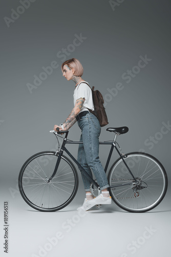 side view of beautiful tattooed girl with backpack riding bicycle on grey