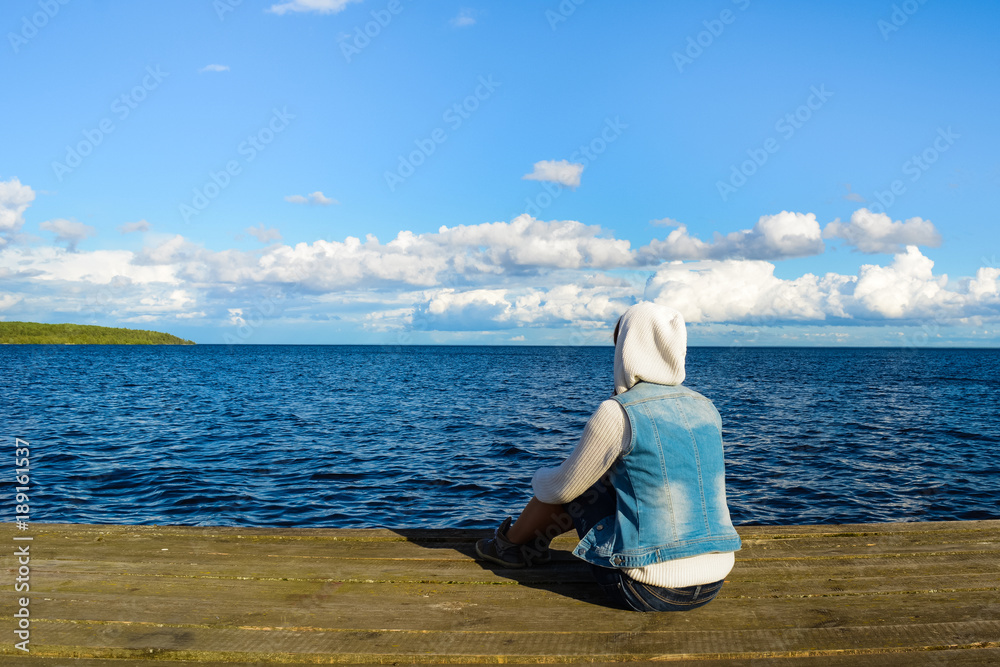 Lonely young woman sitting on wooden mooring near lake and looking on nature, relaxation concept