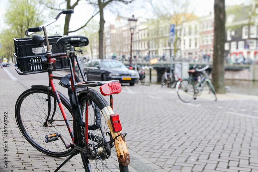 Bicycle parked on the street in the foreground with a typical canal and architecture of Amsterdam, Netherlands slightly out focus on the background during Spring time in the Dutch capital. Bicycle parked on the street in the foreground with a typical canal and architecture of Amsterdam, Netherlands slightly out focus on the background during Spring time in the Dutch capital.