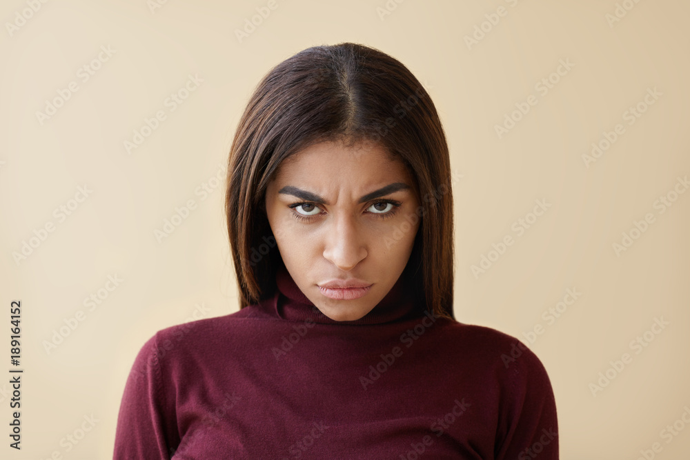 Fototapeta premium Picture of evil young dark skinned brunette woman having sinister appearance, staring at camera from under her frowned eyebrows, grabbing lips furiously, her look full of anger and irritation