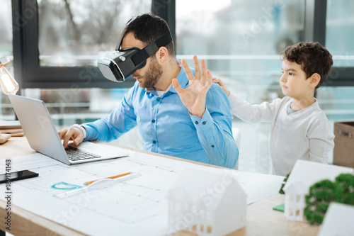 Wait a second. Bristled young architect in a VR headset working on a project and raising his hand as if asking his son touching his back not to distract him