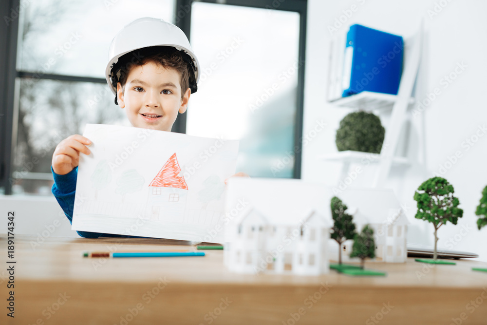 Talented artist. Cute little boy sitting at the table in the architects office and showing a picture of a house, having drawn it by himself Talented artist. Cute little boy sitting at the table in the architects office and showing a picture of a house, having drawn it by himself